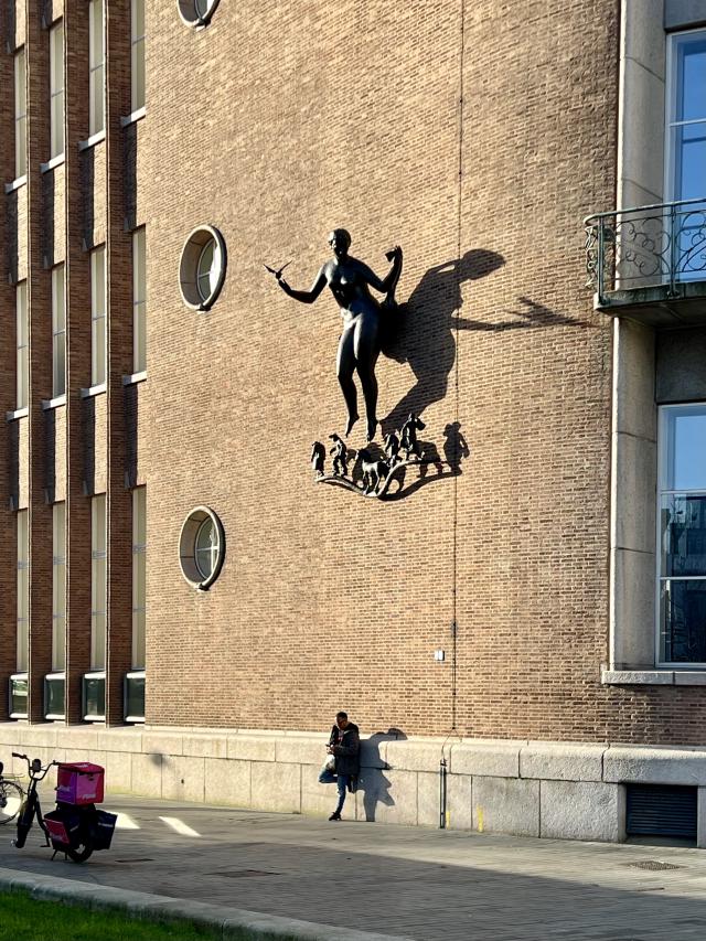 Een man staat tegen een monumentaal gebouw geleund, terwijl de schaduwen over het gebouw trekken