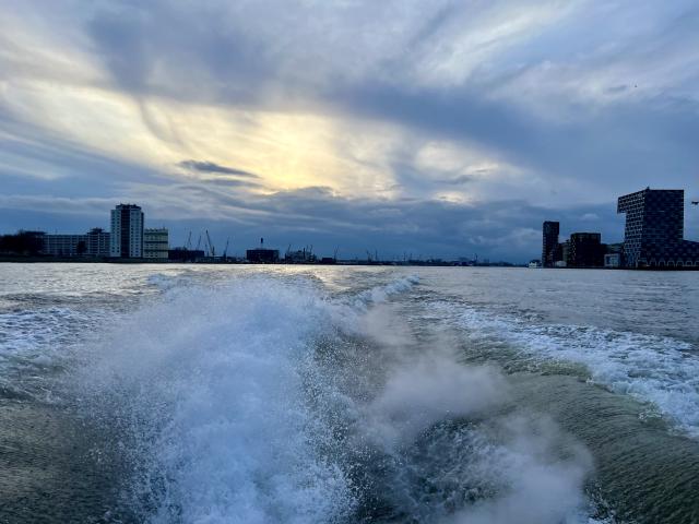 Boot vaart over water met golven, richting een stad met hoge gebouwen onder een bewolkte hemel bij zonsondergang.