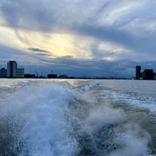Boot vaart over water met golven, richting een stad met hoge gebouwen onder een bewolkte hemel bij zonsondergang.