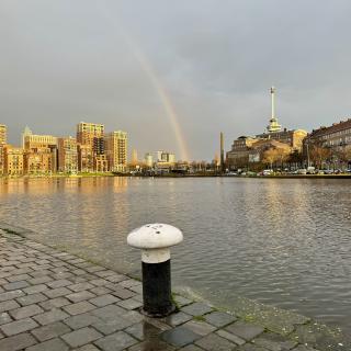 Een regenboog boven een gracht met moderne gebouwen en een klokkentoren in de verte, onder een grijze lucht.