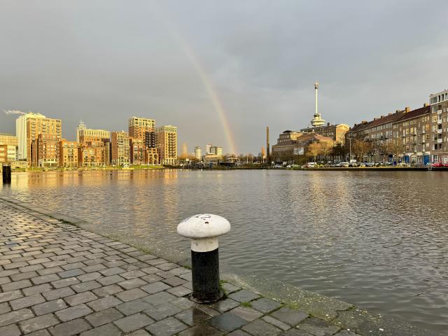 Een regenboog boven een gracht met moderne gebouwen en een klokkentoren in de verte, onder een grijze lucht.