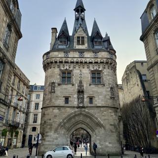 Middeleeuwse poort met torens in Bordeaux, omringd door gebouwen en mensen. Een witte auto staat ervoor op een geplaveide straat.