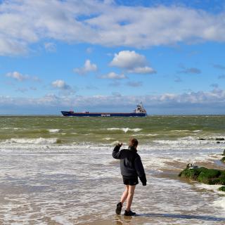 Een persoon in een zwarte jas staat op een strand met golven en een groot schip in de verte onder een blauwe lucht met wolken.