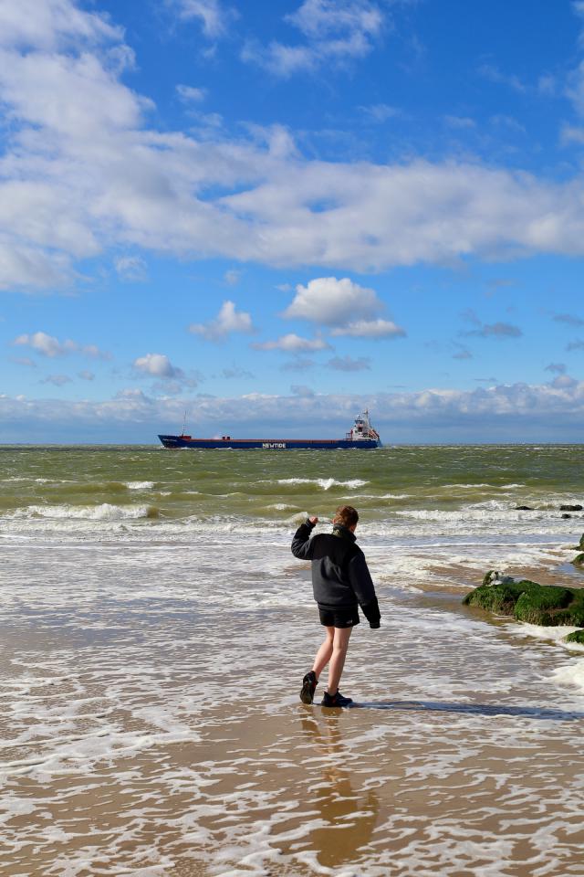 Een persoon in een zwarte jas staat op een strand met golven en een groot schip in de verte onder een blauwe lucht met wolken.