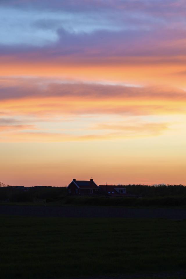 Silhouet van een huis tegen een kleurrijke zonsondergang met oranje, roze en blauwe tinten boven een groen veld.