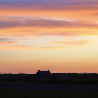 Silhouet van een huis tegen een kleurrijke zonsondergang met oranje, roze en blauwe tinten boven een groen veld.