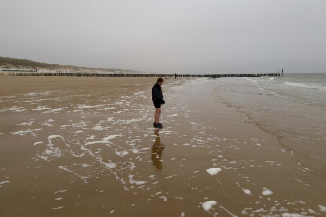 Een persoon staat op een nat strand met een pier in de verte onder een grijze lucht.