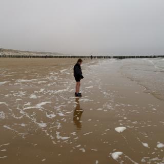 Een persoon staat op een nat strand met een pier in de verte onder een grijze lucht.