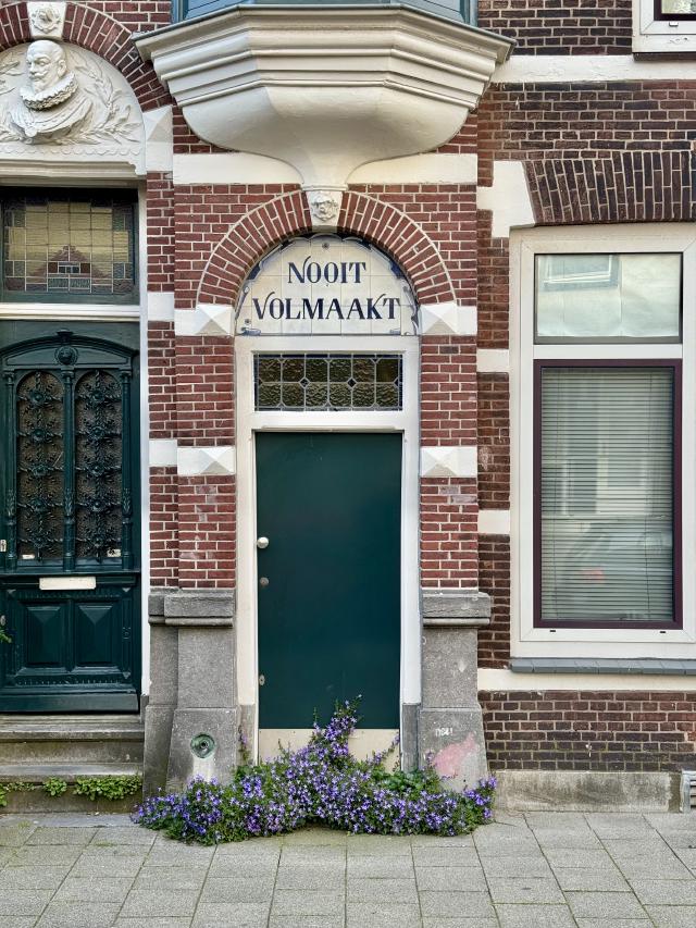 Rode bakstenen gevel met groene deur en decoratieve boog. Boven de deur staat NOOIT VOLMAAKT. Een buste en paarse bloemen voegen charme toe.