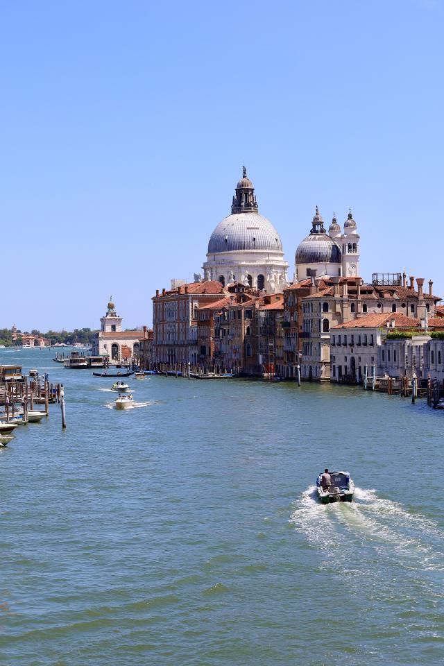 Een boot vaart over een kanaal in Venetië met de Basilica di Santa Maria della Salute op de achtergrond onder een blauwe hemel.