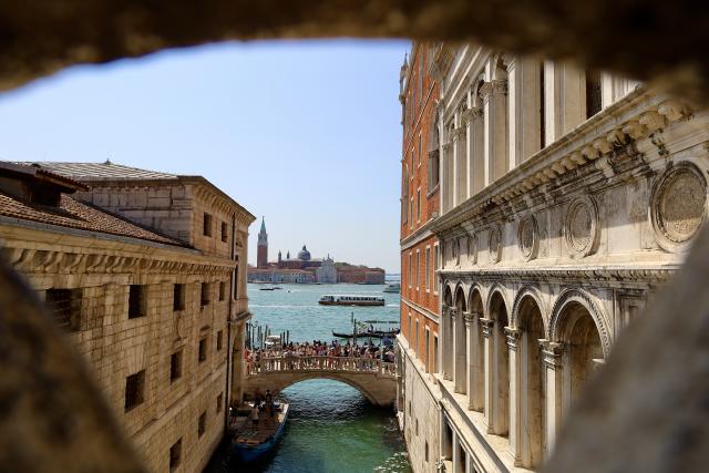Uitzicht door een boog op een kanaal in Venetië met een brug en historische gebouwen. In de verte zie je de San Giorgio Maggiore kerk.
