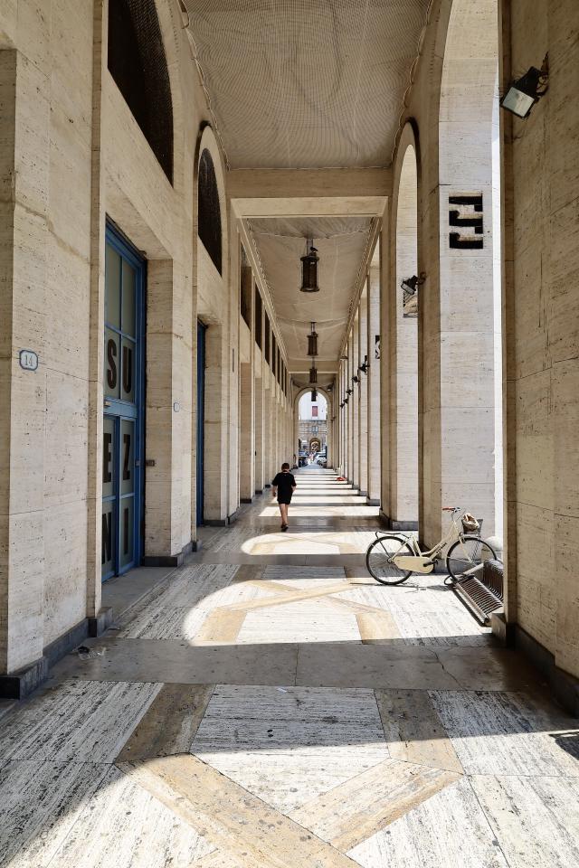 Een lange, overdekte gang met bogen en blauwe deuren, verlicht door hanglampen. Een persoon loopt naar voren en een fiets staat tegen de muur.