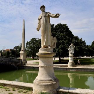 Standbeeld van een man op een sokkel bij een vijver met een obelisk en bomen in de achtergrond.