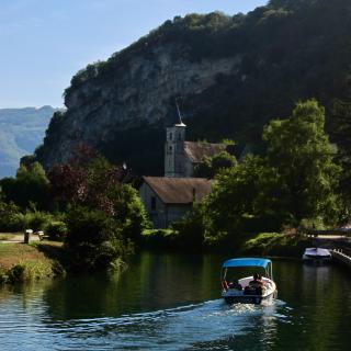Een bootje vaart over een kalme rivier langs een schilderachtig dorpje met een kerkje en een steile rotswand, omgeven door groen en bomen onder een heldere blauwe lucht.
