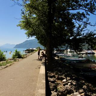 Een pad langs een meer met boten, mensen die ontspannen en bergen in de verte onder een heldere blauwe lucht.
