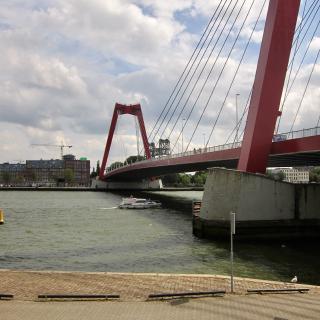 Rode kabelbrug over een rivier met een boot eronder, onder een bewolkte hemel.