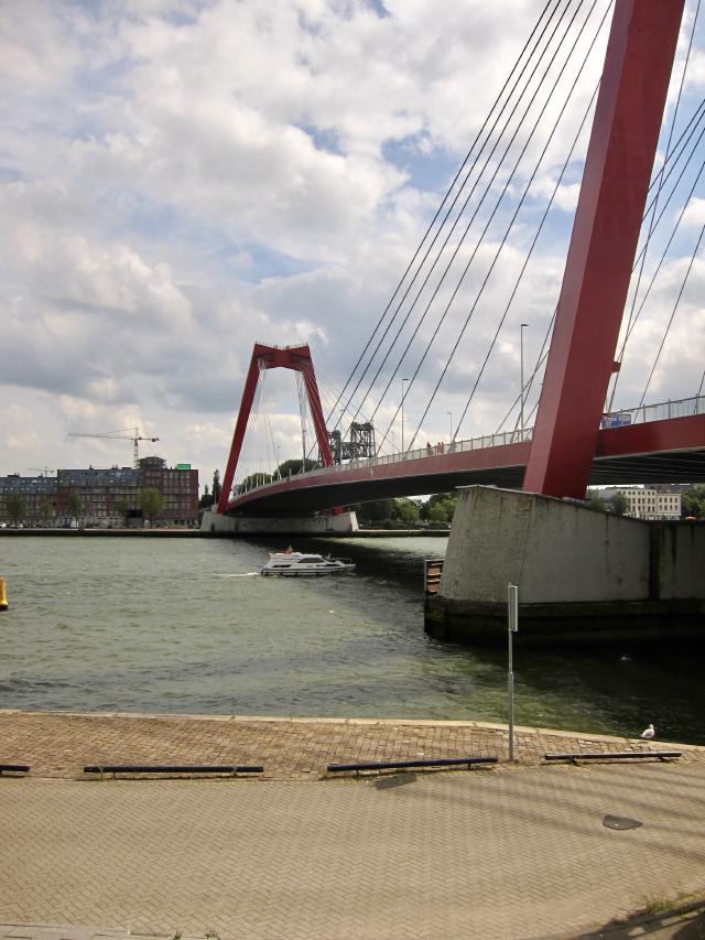 Rode kabelbrug over een rivier met een boot eronder, onder een bewolkte hemel.