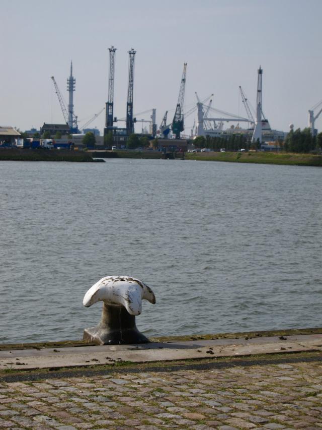 Witte scheepsanker bij een waterkant met kranen en gebouwen in de verte onder een blauwe lucht.