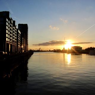 Zonsondergang boven een rivier met moderne gebouwen aan de linkerkant en een heldere hemel met een paar wolken.