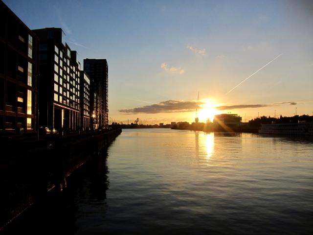 Zonsondergang boven een rivier met moderne gebouwen aan de linkerkant en een heldere hemel met een paar wolken.