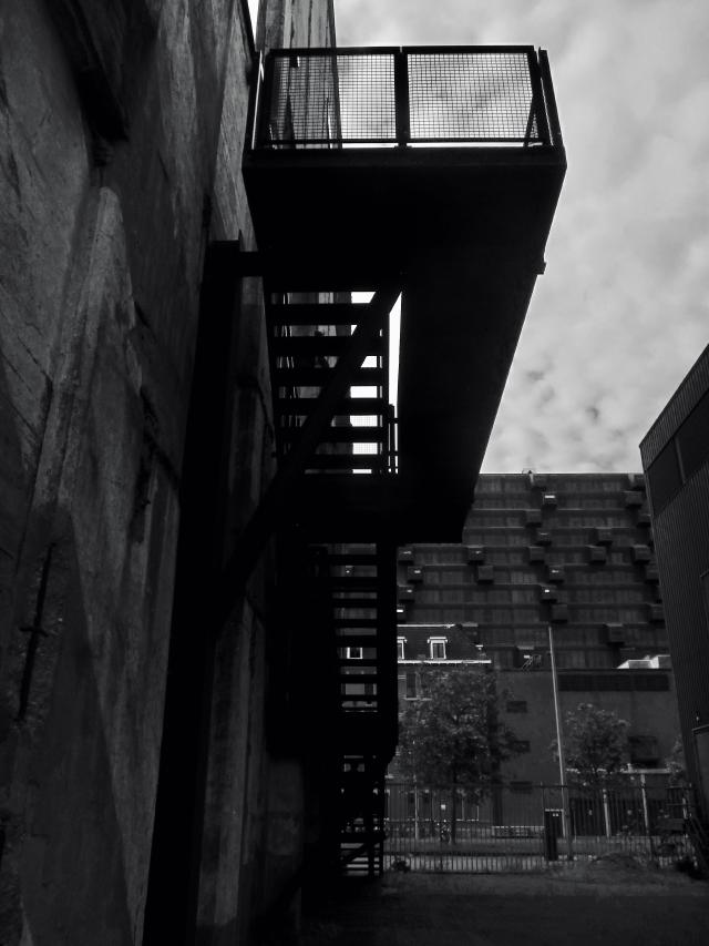 Zwart-wit foto van een moderne loopbrug tussen twee gebouwen, met een mesh railing en een donkere lucht.