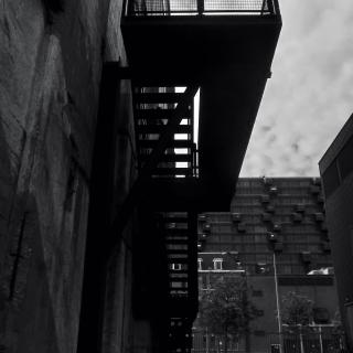 Zwart-wit foto van een moderne loopbrug tussen twee gebouwen, met een mesh railing en een donkere lucht.