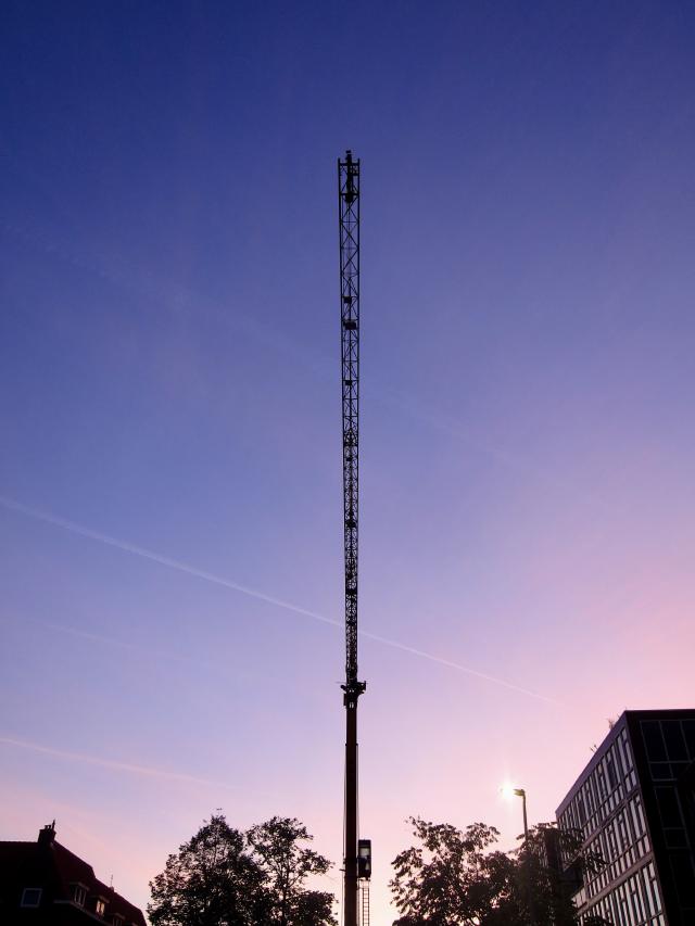 Een hoge bouwkraan tegen een blauwe en roze lucht, met bomen en een gebouw in de voorgrond.