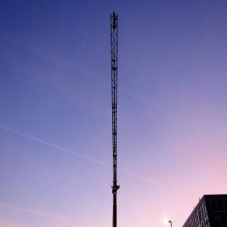 Een hoge bouwkraan tegen een blauwe en roze lucht, met bomen en een gebouw in de voorgrond.