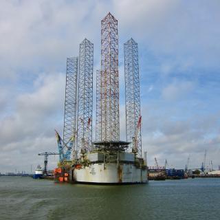 Een offshore booreiland met vier hoge torens in een haven, omgeven door water en kranen onder een bewolkte hemel.