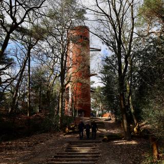 Drie mensen lopen op een pad naar een rode toren in een bos met kale bomen en bladeren op de grond.