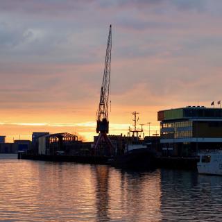 Silhouet van een haven met een grote kraan en schepen bij zonsondergang, reflecterend in het water.