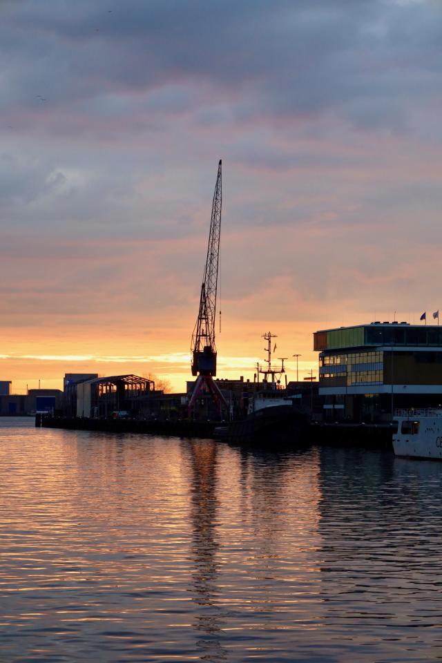 Silhouet van een haven met een grote kraan en schepen bij zonsondergang, reflecterend in het water.