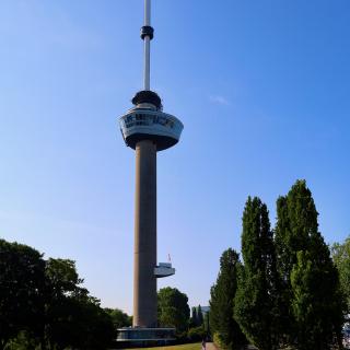 Een hoge, slanke toren met een ronde top en een antenne, omgeven door groen gras en bomen onder een heldere blauwe lucht.