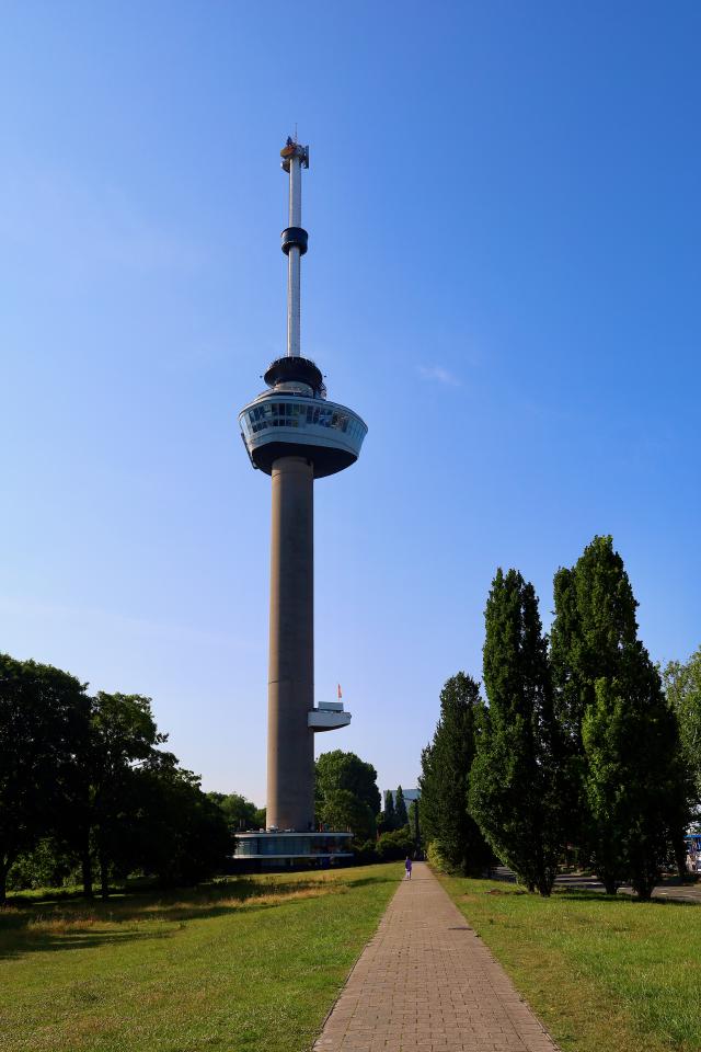 Een hoge, slanke toren met een ronde top en een antenne, omgeven door groen gras en bomen onder een heldere blauwe lucht.