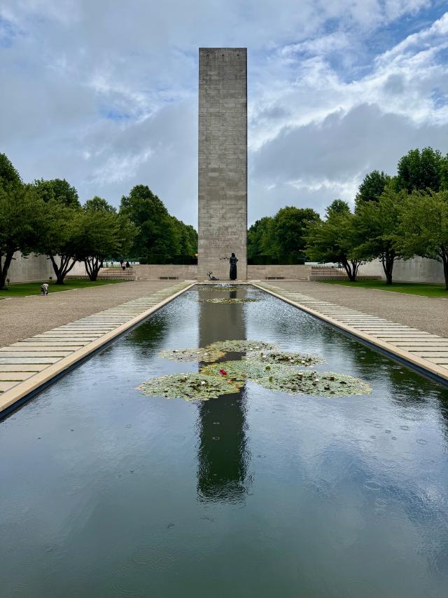 Een hoge stenen obelisk in een rechthoekige vijver met waterlelies, omgeven door bomen en een grijze lucht.