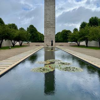 Een hoge stenen obelisk in een rechthoekige vijver met waterlelies, omgeven door bomen en een grijze lucht.