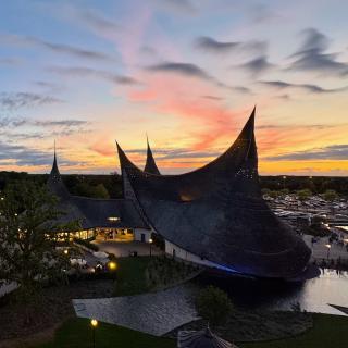Een modern gebouw met spitsvormige daken bij zonsondergang, omgeven door water en groen, met een parkeerplaats in de verte.