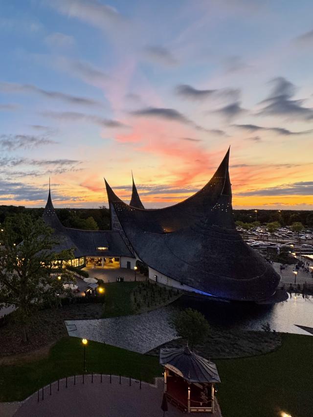 Een modern gebouw met spitsvormige daken bij zonsondergang, omgeven door water en groen, met een parkeerplaats in de verte.