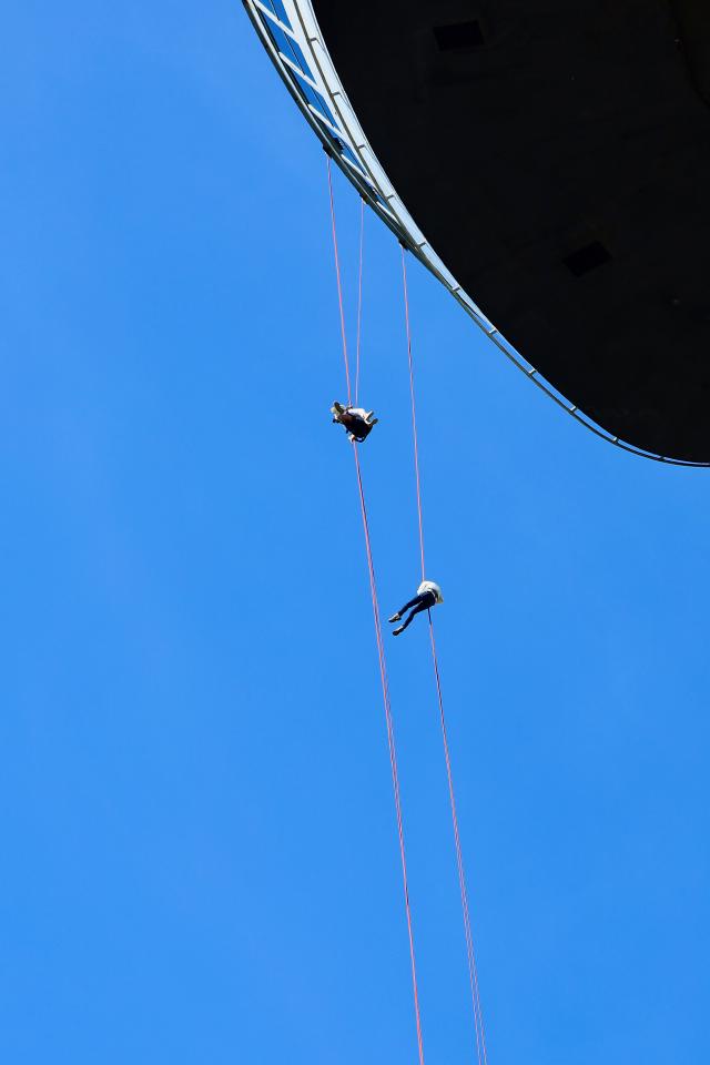 Twee mensen hangen aan touwen onder een blauwe lucht, dicht bij een groot zwart object.