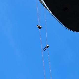 Twee mensen hangen aan touwen onder een blauwe lucht, dicht bij een groot zwart object.