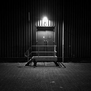 Donkere nacht met een verlichte deur in een metalen wand, omgeven door een leeg asfaltplein.