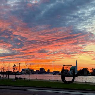 Een kleurrijke zonsondergang boven een rivier met een stadssilhouet en een groot zwart kunstwerk op de voorgrond.