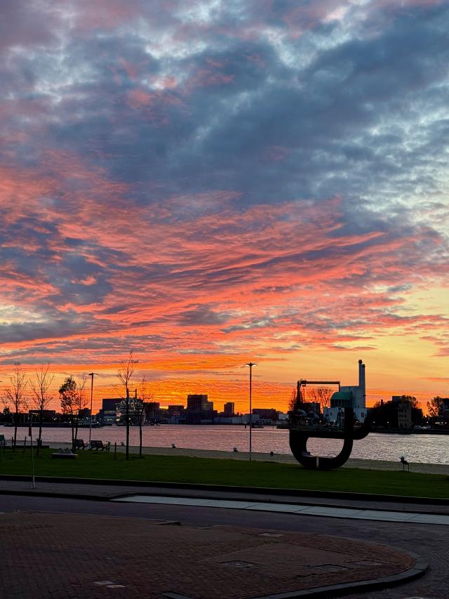 Een kleurrijke zonsondergang boven een rivier met een stadssilhouet en een groot zwart kunstwerk op de voorgrond.