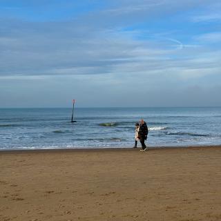 Twee mensen wandelen op een zandstrand met een blauwe lucht en een rode paal in de zee.