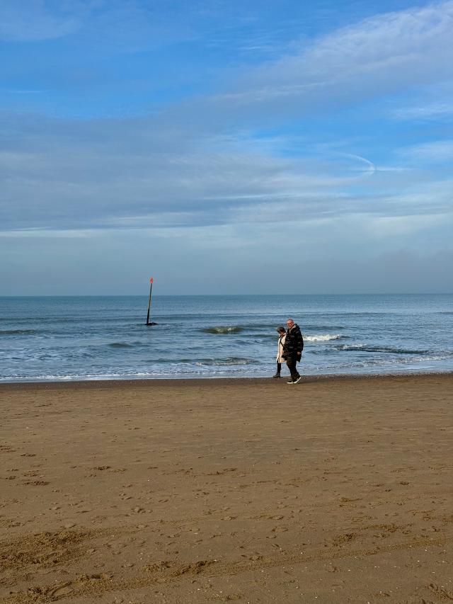 Twee mensen wandelen op een zandstrand met een blauwe lucht en een rode paal in de zee.