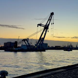 Silhouet van grote kranen in een haven bij zonsondergang, met water en een stenen kade in de voorgrond.